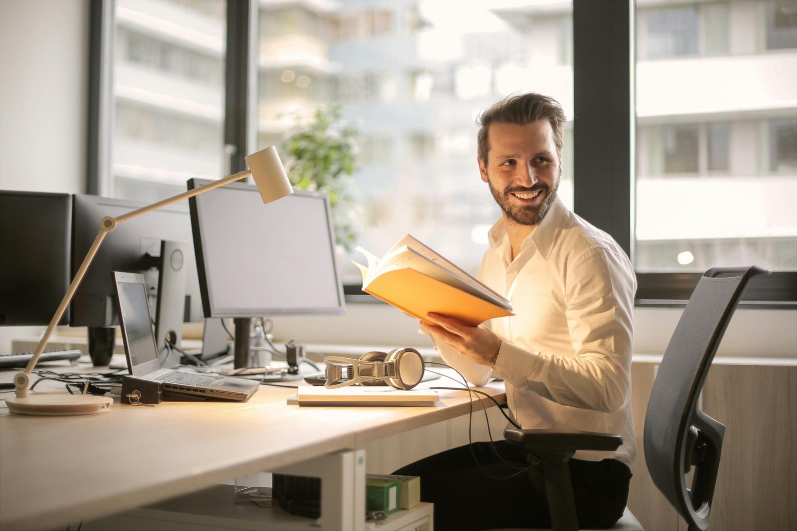 A man smiling and holding a book while sitting at a desk in a modern office, with two computers and a desk lamp visible in the workspace.