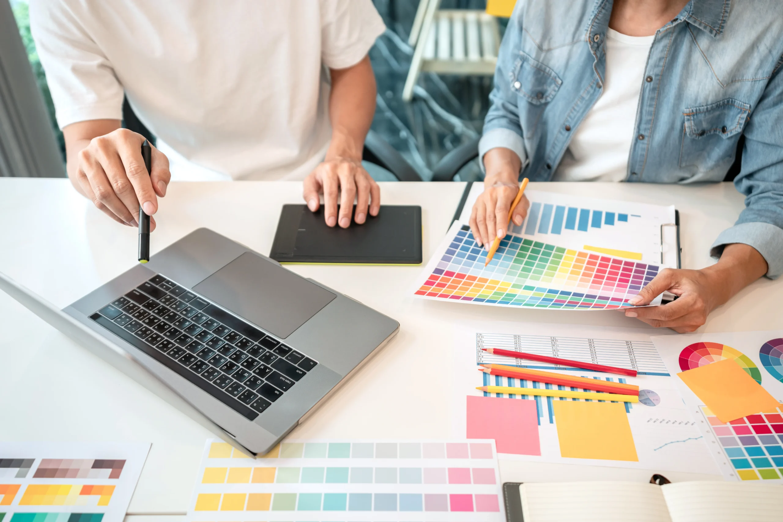 Two people working collaboratively at a desk, one holding a stylus over a laptop and the other examining a color palette and design charts.